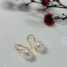 Load image into Gallery viewer, Pearl earrings with gold hooks on a white surface with pink flowers in the background
